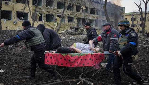 Emergency employees and volunteers carry an injured woman, heavily pregnant, on a  stretcher past the smoldering wreckage of Mariupol’s hospital that was damaged by  shelling on Wednesday, March 9, 2022. Her face is pale, one hand cradles her belly in  a protective gesture. Every window on that side of the building appears to be blown out  and wreckage litters the ground. (Tim Lister, Laura Smith-Spark, Olga Voitovych and Rob  Picheta: CNN,Kyiv.March 10, 2022.  Photo by Evgeniy Maloletka).